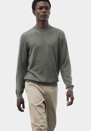 Jonge man loopt, draagt een grijze trui met lange mouwen en beige broek tegen een effen lichte achtergrond.
