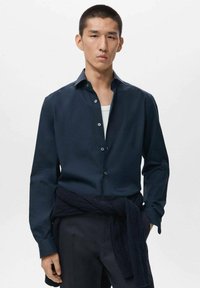 Camisa azul marino de botones, con cuello extendido, mangas largas y textura suave. Combinada con pantalones oscuros y un suéter azul marino atado a la cintura.