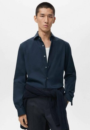 Camisa azul marino de botones, con cuello extendido, mangas largas y textura suave. Combinada con pantalones oscuros y un suéter azul marino atado a la cintura.