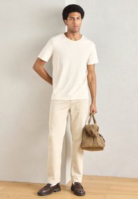 Μπεζ βαμβακερό T-shirt, ανοιχτό μπεζ παντελόνι κοτλέ, σκούρα καφέ παπούτσια και καφέ δερμάτινη τσάντα με χερούλια. Το μοντέλο στέκεται μπροστά σε ανοιχτόχρωμο τοίχο.