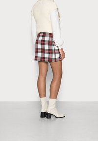 Jupe mini à carreaux en blanc, rouge et noir avec une coupe droite, assortie à un gros pull crème et des bottines blanches avec un talon noir.