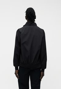 Άτομο με σκουρόχρωμο δέρμα και κοντά dreadlocks, ντυμένο με μαύρο μπουφάν και μαύρο παντελόνι, με την πλάτη γυρισμένη προς ένα απλό λευκό φόντο.