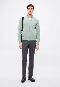 Ανοιχτό πράσινο σακάκι με φερμουάρ και μακριά μανίκια, συνδυασμένο με γκρι παντελόνια σε ραμμένη γραμμή και μαύρα δερμάτινα παπούτσια, με διακριτικό λογότυπο.