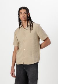 Lyse beige kortærmet skjorte lavet af tekstureret stof, med en knappet front og en afslappet pasform, parret med mørke jeans.