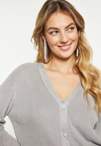 Cardigan in maglia argento con scollo a V, caratterizzato da due bottoni decorativi e una finitura testurizzata. Maniche lunghe e oltre a un lieve luccichio metallico.