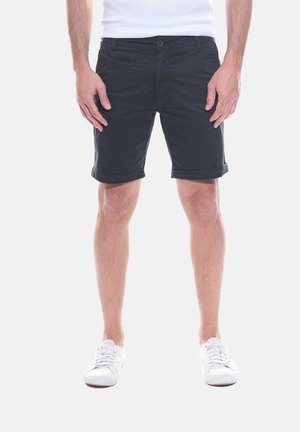 Homme portant un short noir retroussé, des baskets blanches et une chemise blanche, debout les mains détendues le long du corps sur un fond blanc uni.