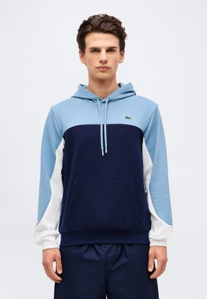 Junger Mann trägt einen blau-, marine- und weißfarbenen Colorblock-Hoodie mit Fronttasche und Lacoste-Logo auf der Brust, steht vor schlichtem Hintergrund.