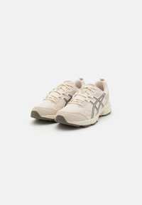 Ténis de desporto bege com tecido em malha, design com atacadores e detalhes cinzentos. Apresenta uma sola almofadada e uma sola texturizada para tração.