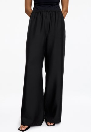 Femme portant un pantalon noir taille haute et coupe large, associé à un top noir sans manches ainsi qu’à des sandales noires à bout carré.