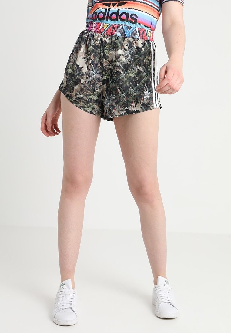 Adidas-Shorts mit einem schwarzen Palmenblattmuster, elastischem Bund und weißen seitlichen Streifen. Kombiniert mit einem bunten gestreiften Oberteil und weißen Turnschuhen.
