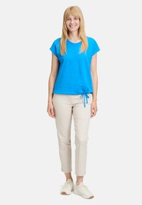 Niebieska bluzka z krótkim rękawem o fakturowanym wzorze, z detalem w postaci boku związania, zestawiona z beżowymi spodniami slim fit oraz jasnymi trampkami.