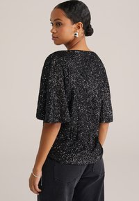 Top nero con paillettes, maniche corte e scollatura ampia, caratterizzato da una superficie testurizzata e dettagli sul retro, abbinato a pantaloni scuri a vita alta.
