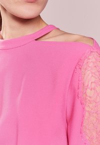 Rosa blus med en slät tygkvalitet, en asymmetrisk urringning och spetsdetalj på axeln, med en texturerad yta och ett fint mönster.