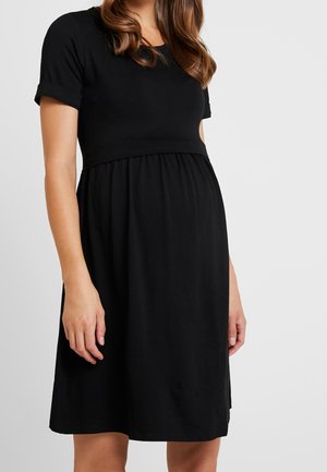 Femme portant une robe de maternité noire mi-longue, à manches courtes et taille cintrée au-dessus du ventre arrondi.