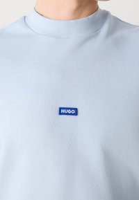 T-shirt bleu clair à col rond avec un petit patch rectangulaire bleu portant l'inscription "HUGO" centré sur la poitrine.