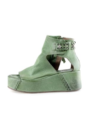Zielony zamszowy sandał na koturnie z odkrytymi palcami, wycięciami po bokach, paskiem wokół kostki z metalowymi ćwiekami oraz ozdobnym przeszyciem na platformie.