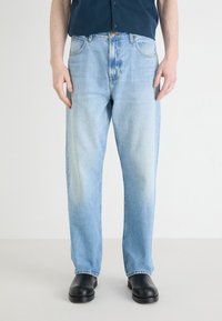 Jeans in denim azzurro chiaro con taglio dritto e lavaggio sbiadito. Presentano cinque tasche e una chiusura con bottone. Indossati con scarpe nere.