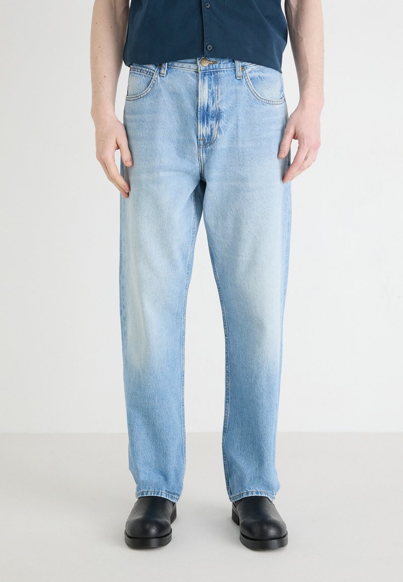 Jeans in denim azzurro chiaro con taglio dritto e lavaggio sbiadito. Presentano cinque tasche e una chiusura con bottone. Indossati con scarpe nere.