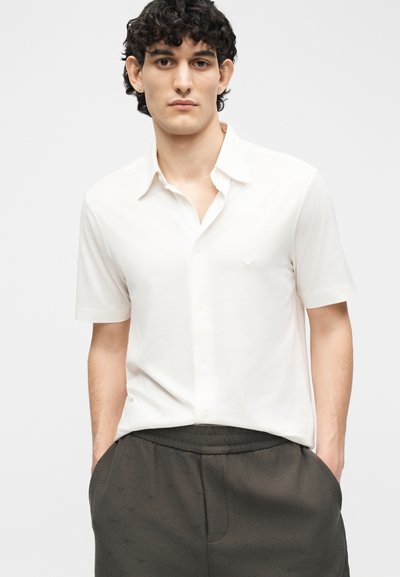 Νέος άνδρας με σγουρά σκούρα μαλλιά, φοράει άσπρο πουκάμισο με κοντά μανίκια και σκούρο παντελόνι, με τα χέρια στις τσέπες, σε απλό φωτεινό φόντο.