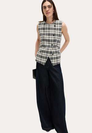 Vrouw draagt een mouwloos zwart-wit geruit vest met knopen en een wijde donkere spijkerbroek, staat met handen in de zakken.