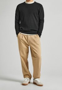 Man som bär svart tröja, beige veckade byxor och vita sneakers med gummiklädda sulor, stående med händerna i fickorna.