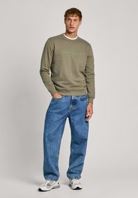 Sudadera verde oliva con logo bordado en verde, combinada con jeans claros y holgados. El modelo lleva zapatillas blancas.