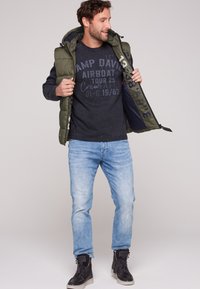 Gilet verde con cappuccio, maglione blu navy con stampa, jeans in denim azzurro e sneakers nere alte. Outfit casual.