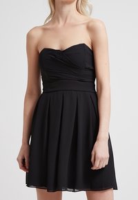 Zwarte strapless jurk met geplooide rok, gemaakt van lichtgewicht stof. Heeft een aansluitende lijfje met een gedraaide ontwerp bij de buste.