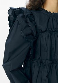 Blouse bleu marine en tissu léger avec des volants en forme de vagues sur les épaules et une taille élastiquée pour un ajustement décontracté.