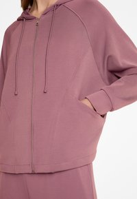 Hellrosa Zip-Hoodie mit Kapuze, vorderen Taschen und Raglanärmeln. Glatte Textur, lockere Passform und metallischer Reißverschlussdetails.