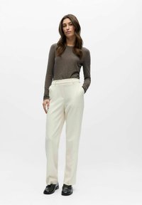 Kremfargede, skreddersydde bukser med sidelommer og en glatt tekstur, kombinert med en tettsittende brun genser med lange ermer. Sorte sko fullfører antrekket.