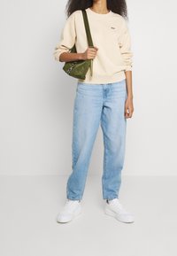 Sweat-shirt crème avec un petit logo, jean baggy bleu clair, baskets blanches, et sac bandoulière vert olive avec un accent en chaîne.