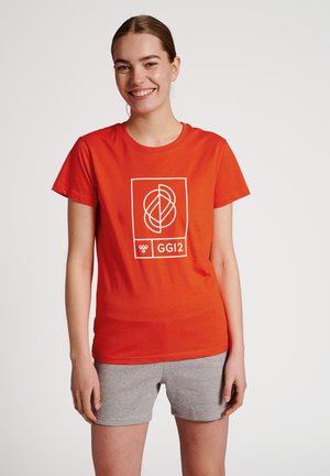 Smilende kvinde med bundet hår iført en rød T-shirt med hvidt geometrisk logo og teksten "GG12", kombineret med grå shorts.
