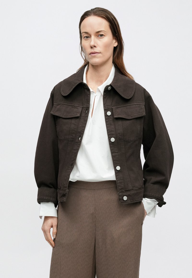 Veste en denim marron avec grand col, deux poches poitrine et fermetures à pression, associée à un chemisier blanc et un pantalon marron texturé.