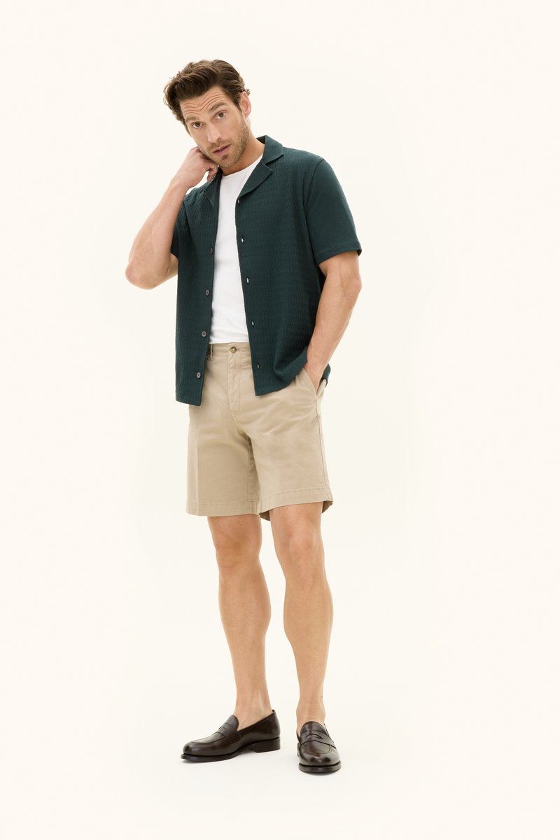 Mann, der ein dunkelgrünes Kurzarmhemd über einem weißen T-Shirt, beigen Shorts und dunkelbraunen Loafers trägt, steht mit einer Hand in der Tasche.