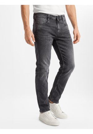 Mann trägt ausgewaschene, schwarze Slim-Fit-Jeans und weiße Turnschuhe, steht vor einem schlichten weißen Hintergrund.