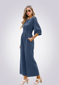 Marineblauwe jumpsuit met korte mouwen, smocktail en zijzakken. Lichtgewicht stof met een gestructureerde afwerking. Draag met blokhak sandalen.