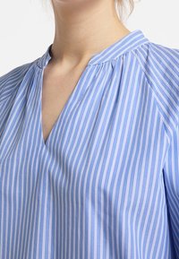 Ljusblå, randig blus med v-ringning, plisserade detaljer vid axlarna och mjukt, lätt material.
