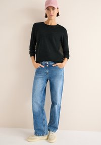 Schwarzes Langarmshirt, hellblaue Schlagjeans und beigefarbene Sneakers. Das Model trägt eine rosa Cordmütze. Glattes Material, lockere Passform.