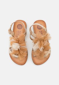 Dziecięce sandały ze skóry w kolorze brązowym, z regulowanymi paskami w okolicy kostki i dużymi beżowymi oraz brązowymi dekoracjami z tiulu w kształcie kwiatów z przodu.