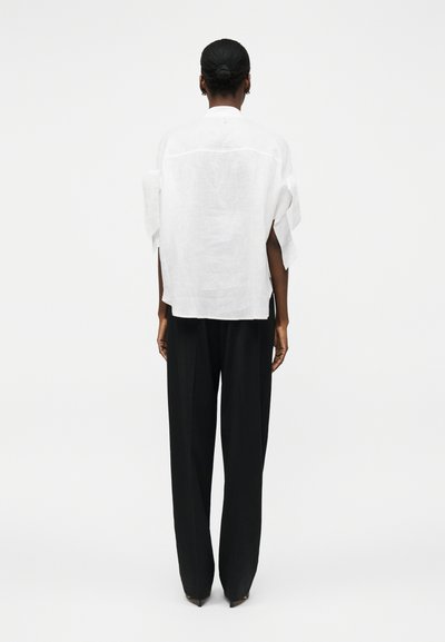 Personne à la peau foncée et aux cheveux en chignon porte une chemise blanche ample et un pantalon noir à jambes larges, debout de dos sur fond blanc.