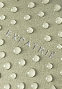 Materiale verde chiaro texture con gocce in rilievo, con la scritta in rilievo argento "EXPATRIÉ" in mostra.