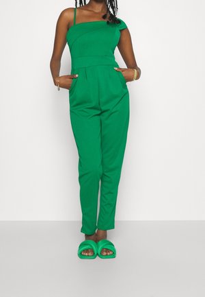 Kobieta w jaskrawozielonym kombinezonie na jedno ramię i pasujących zielonych sandałach wsuwanych, stojąca z rękami w kieszeniach.