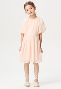 Pfirsichfarbendes, gefaltetes Kleid mit kurzen Puffärmeln, gerafftem Taillenbereich und ausgestelltem Rock. Kombiniert mit neutralen Espadrilles und Bandakzenten an den Knöcheln.