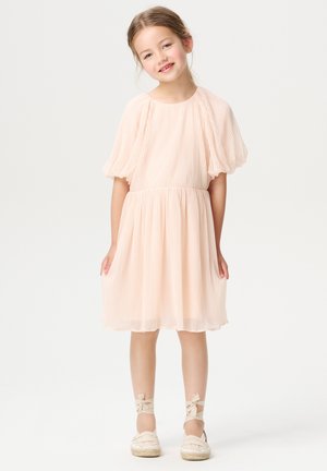 Pfirsichfarbendes, gefaltetes Kleid mit kurzen Puffärmeln, gerafftem Taillenbereich und ausgestelltem Rock. Kombiniert mit neutralen Espadrilles und Bandakzenten an den Knöcheln.