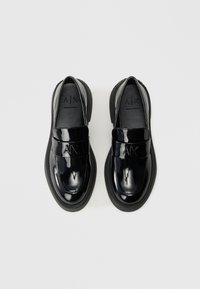 Crni lakirani slip-on mokasini s okruglim vrhom, debelim đonom, utisnutim logotipom na trakici i glatkom teksturom. Gornji pogled na bijeloj pozadini.