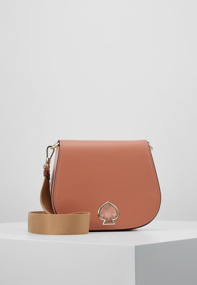 Sac bandoulière en cuir marron avec large sangle beige et logo pique doré sur un piédestal blanc sur fond gris.