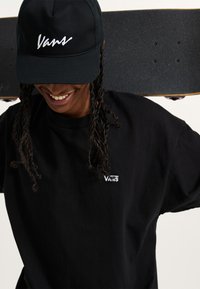 Schwarzes Baumwoll-T-Shirt mit weißem "Vans"-Logo, getragen von einer Person, die ein Skateboard hält. Schwarze Kappe mit weißem besticktem Logo.