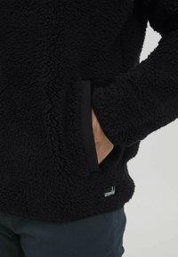 Schwarzer Fleece-Pullover mit einer weichen, strukturierten Oberfläche, ausgestattet mit seitlichen Taschen und schwarzem Stoffbesatz. Logopatch am unteren Saum.