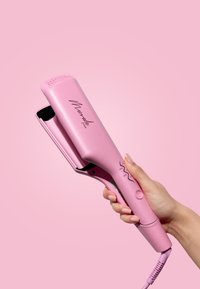 Plancha de pelo rosa con un acabado suave, bordes redondeados y una pantalla digital de temperatura. Sostenida por una mano contra un fondo rosa.